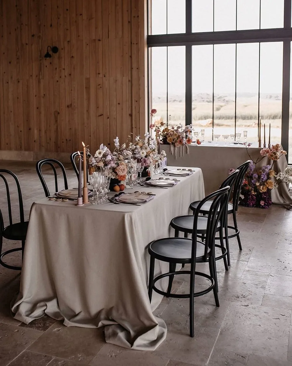A long table dressed in linen with candles, flowers and black bistro chairs, in front of tall windows