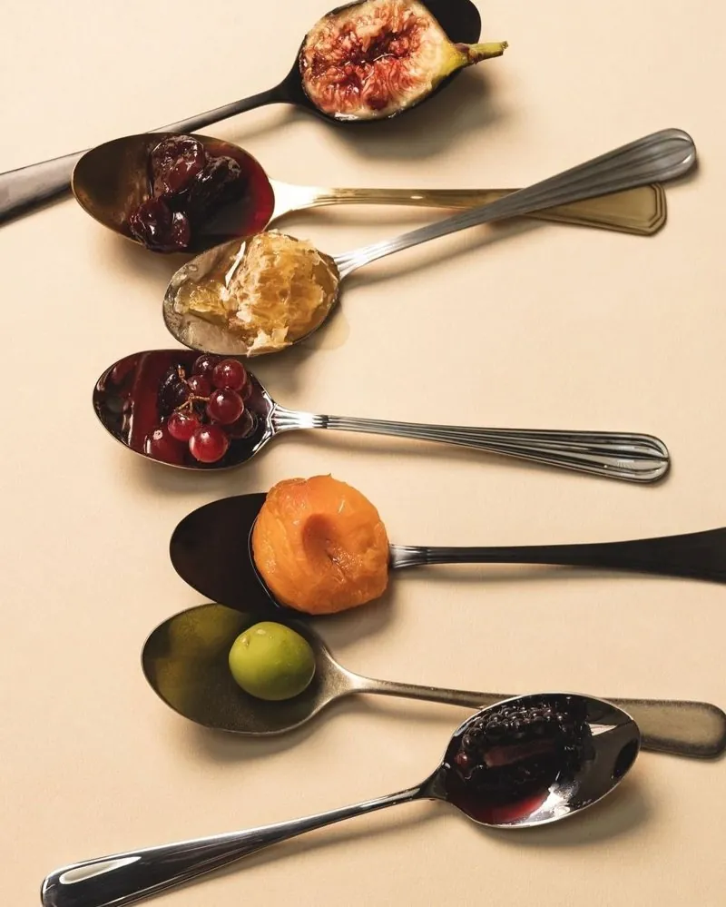 Seven spoons holding a fig, raisins, honeycomb, grapes, an apricot, an olive and a blackberry